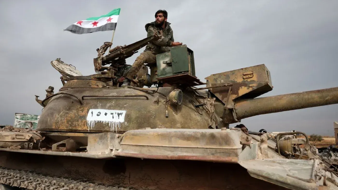 مقاتل من قوات المعارضة السورية يجلس على دبابة قديمة، حاملاً علم الثورة السورية، في سياق الاشتباكات المستمرة في الرقة.