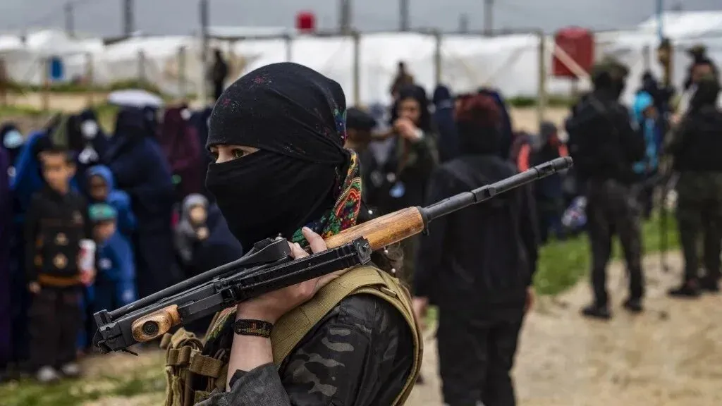 امرأة مسلحة ترتدي غطاء رأس أسود، تقف في مقدمة مجموعة من الأشخاص في مخيم، مما يعكس الأوضاع الأمنية في شمال سوريا بعد الاتفاقات الأخيرة.