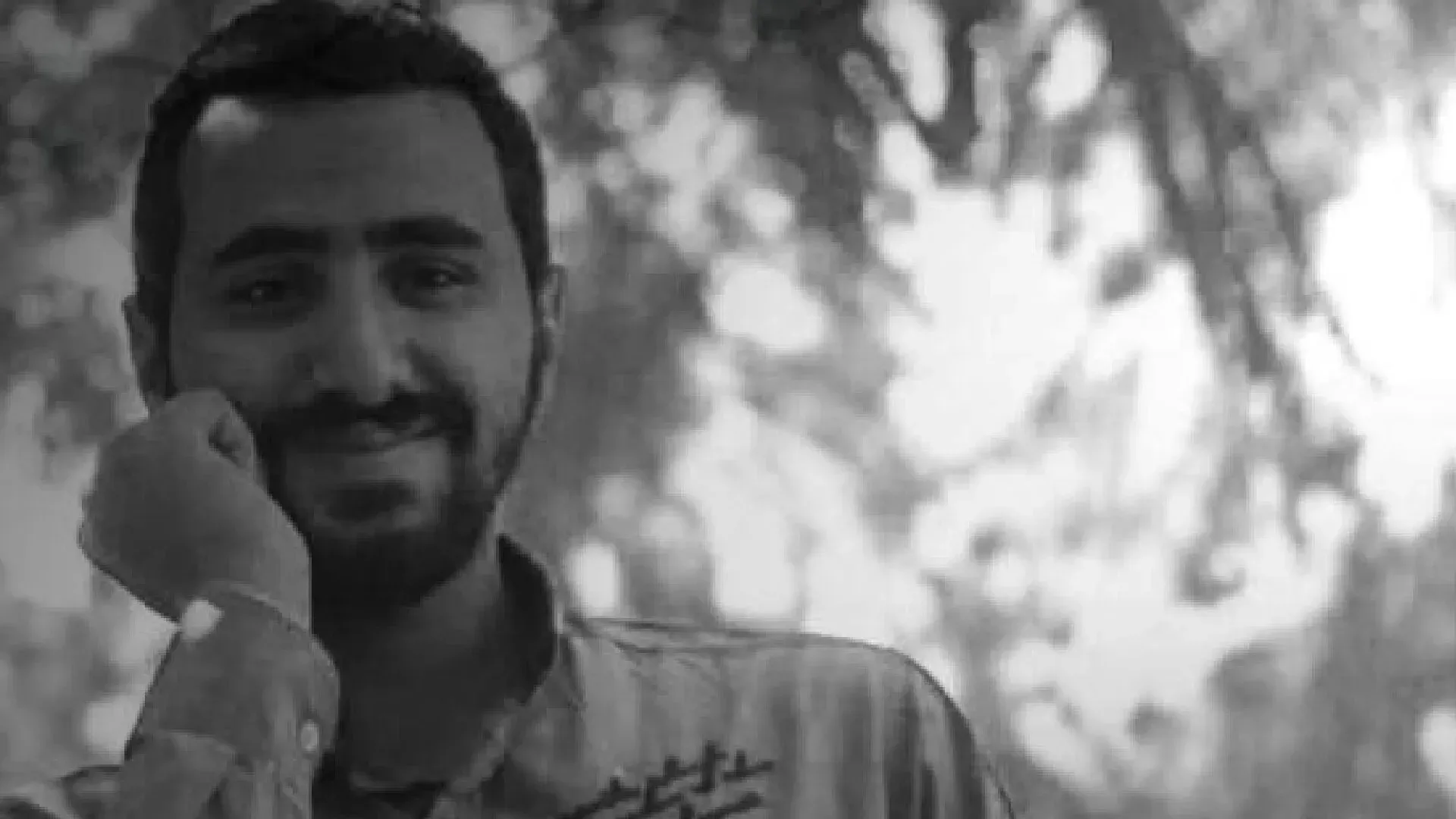 شاب مبتسم يرتدي قميصًا رماديًا، يُظهر صورة شخصية في بيئة طبيعية، تعكس الظروف الإنسانية الصعبة للمتظاهرين في مصر.
