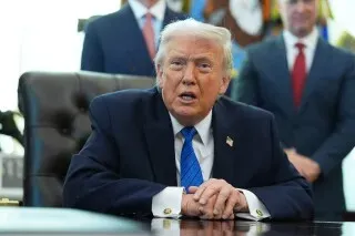 ترامب يجلس خلف مكتب في المكتب البيضاوي، مع تعبير جاد، بينما يظهر خلفه بعض المساعدين. الصورة تعكس الأجواء السياسية المتوترة.