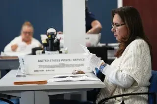 امرأة ترتدي قفازات بيضاء تعمل في مكتب البريد، تفحص بطاقات الاقتراع. تظهر خلفها موظفات أخريات في بيئة مكتبية.