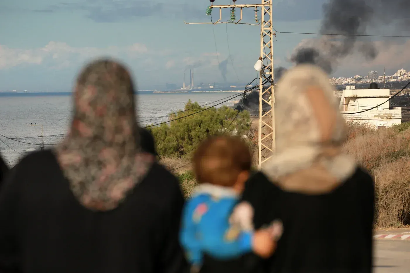 نساء تحملن أطفالهن، يراقبن الدخان يتصاعد من خلف البحر في غزة، مما يعكس الأوضاع المتوترة في المنطقة.