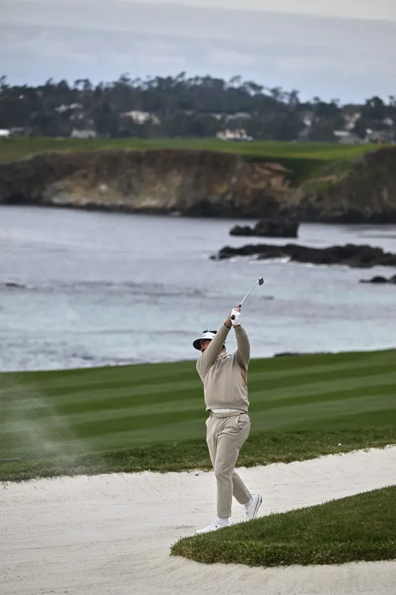 أحد لاعبي الجولف يقوم بضربة من رمال الشاطئ في بطولة AT&T Pebble Beach Pro-Am، مع منظر طبيعي جميل للمحيط وخلفية تلال خضراء.