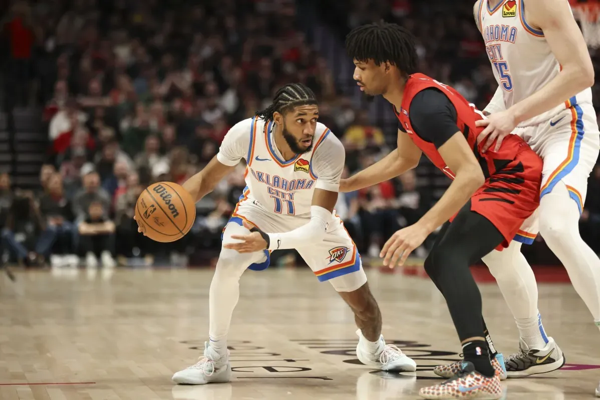 شاي جيلجوس-أليكساندر من أوكلاهوما سيتي يت dribble الكرة أمام لاعب من بورتلاند في مباراة كرة السلة، مع وجود مشجعين في الخلفية.