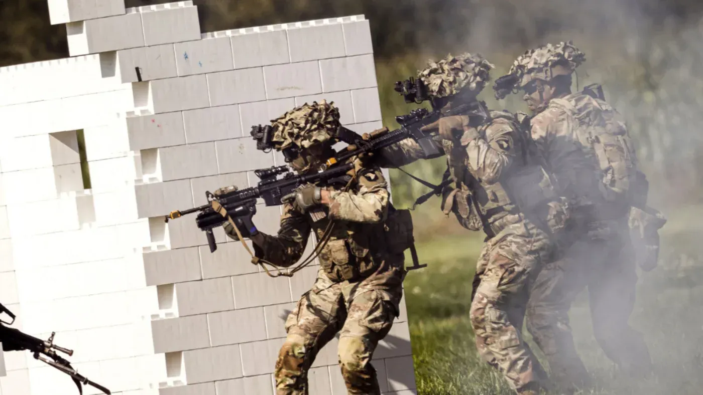 جنود أمريكيون يرتدون زيًا عسكريًا يتقدمون بحذر خلال تدريب عسكري، مع وجود دخان في الخلفية، مما يعكس استعدادهم لعمليات محتملة في الخليج.