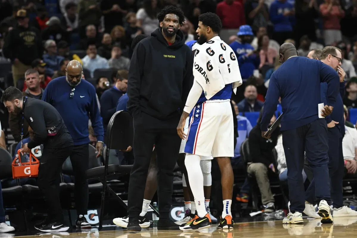 جويل إمبيد يرتدي ملابس رياضية ويتحدث مع زميله في فريق فيلادلفيا 76ers، مع وجود مشجعين في الخلفية، في سياق إصابته الأخيرة.
