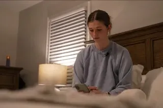 امرأة تجلس على السرير في غرفة نوم مضاءة، تستخدم هاتفها الذكي لمراقبة بيانات نومها، مما يعكس أهمية تتبع النوم في تحسين العادات الصحية.