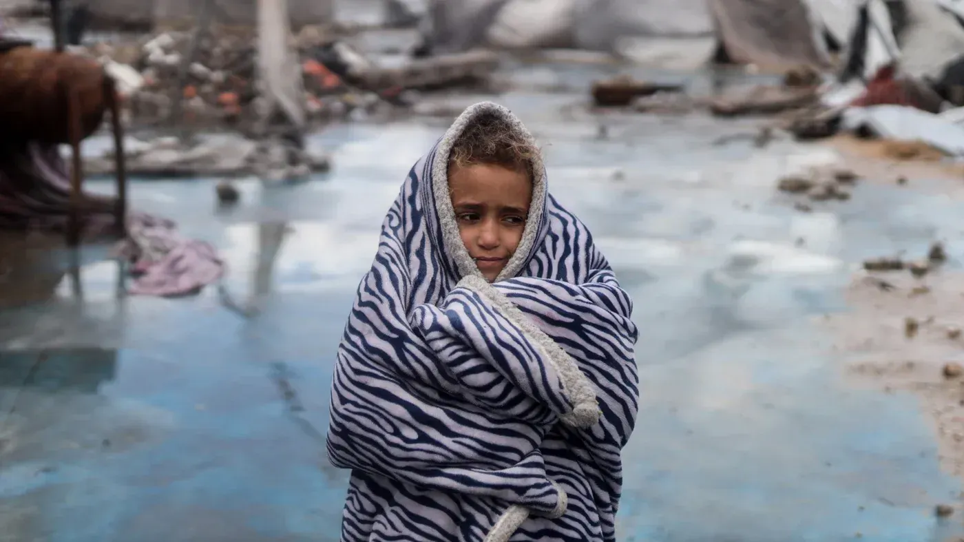 طفل فلسطيني wrapped in a striped blanket، يبدو حزينًا ومرتبكًا وسط أنقاض المنازل المدمرة في غزة، مع تدهور الأوضاع الإنسانية.