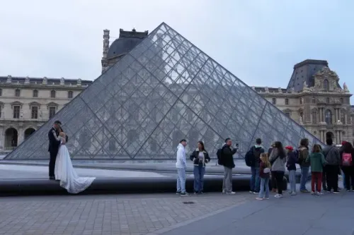 زوجان يرتديان ملابس الزفاف يتصوران أمام الهرم الزجاجي لمتحف اللوفر، مع مجموعة من الزوار في الخلفية.
