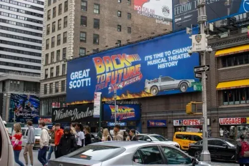 لافتة إعلانية كبيرة لفيلم "العودة إلى المستقبل: الموسيقي" في منطقة ترفيهية مزدحمة، مع مشاة وسيارات تمر في الشارع.