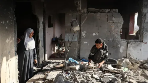 امرأة مسنّة ترتدي حجابًا تقف في غرفة مدمرة بينما يجلس شاب على الأرض وسط الأنقاض، يبحث عن أشياء مهمة في غزة بعد النزاع.