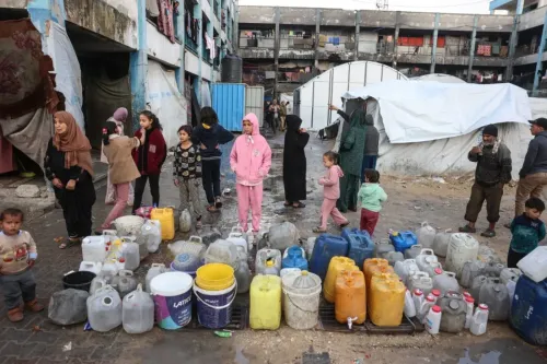 نساء وأطفال يقفون بجوار صف من حاويات المياه في مخيم للاجئين في غزة، مما يعكس الوضع الإنساني الصعب واحتياجات المساعدة الأساسية.