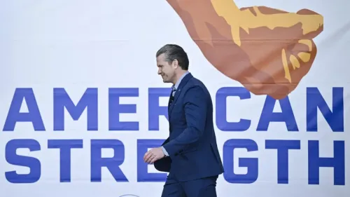 رجل يرتدي بدلة زرقاء يسير بجانب لافتة كبيرة تحمل عبارة "القوة الأمريكية" مع رسم ليد مرفوعة، تعكس روح القوة والهيمنة.