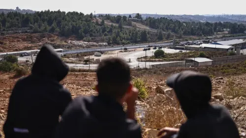 مجموعة من الشباب يجلسون على تلة، يراقبون نقطة تفتيش عسكرية محاطة بالأشجار، مما يعكس التوتر في الضفة الغربية.