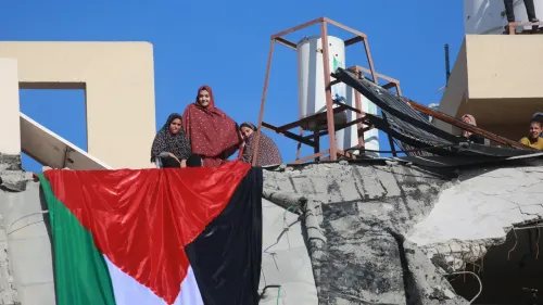 نساء فلسطينيات يقفن على أنقاض مبنى مدمّر، يحملن علم فلسطين، مما يعكس تأثير النزاع المستمر على المجتمع الفلسطيني.