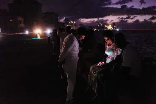 تظهر الصورة مجموعة من الأشخاص يجلسون على كورنيش هافانا ليلاً، مع إضاءة خافتة من المباني القريبة، مما يعكس حالة الهدوء والركود في الحياة الليلية.