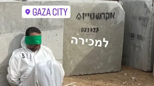 رجل فلسطيني يرتدي بدلة واقية بيضاء، معصوب العينين، مقيد اليدين والقدمين، يجلس بجانب كتل إسمنتية في غزة.