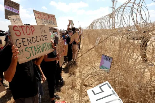 محتجون يحملون لافتات تطالب بوقف المجاعة في غزة، يقفون أمام سياج شائك، معبرة عن معارضة التجويع كتكتيك حربي.