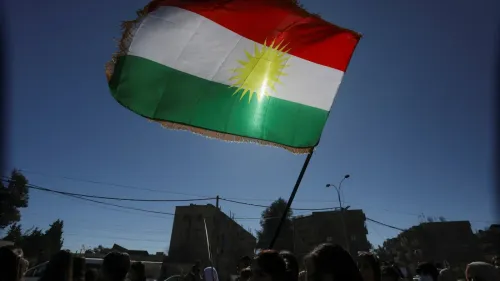 علم كردي يرفرف في الهواء، مع خلفية لمباني في منطقة حضرية، يعكس التوترات السياسية والجهود الكردية ضد النظام الإيراني.