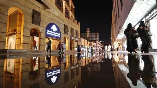 تظهر الصورة مشهدًا ليليًا في مدينة سعودية، حيث يتجول الناس في الشارع، مع انعكاسات واضحة على الأرض المبللة، مما يعكس الحياة الحضرية.