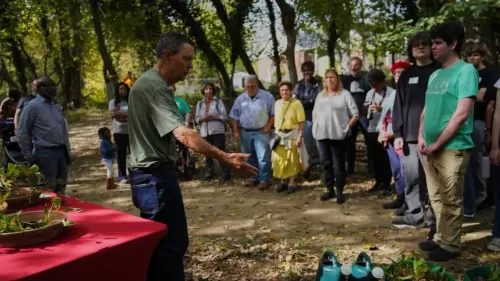 اجتماع مجتمعي في بالتيمور حيث يتحدث رجل أمام جمهور متنوع، مع وجود طاولات مملوءة بالطعام في بيئة طبيعية.