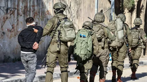 جندي إسرائيلي يقود فلسطينيًا معصوب العينين خلال عملية اعتقال في الضفة الغربية، مع وجود جنود آخرين في الخلفية.