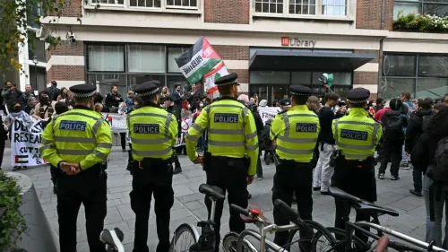تظهر الصورة مجموعة من رجال الشرطة يرتدون زيهم الرسمي، يقفون أمام مكتبة LSE في لندن، بينما يحتشد المتظاهرون خلفهم حاملين لافتات وأعلام فلسطينية، في سياق مسيرة تضامن مع فلسطين.
