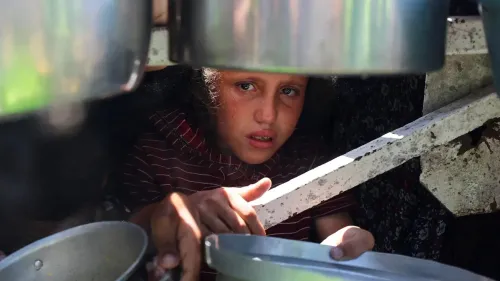طفل فلسطيني يظهر بملامح حزينة وهو يجلس تحت مجموعة من الأواني، يعكس معاناة الجوع وسوء التغذية في غزة.