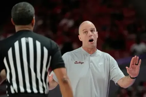 مدرب فريق أوكلاهوما ستيت، ستيف لوتز، يتحدث مع الحكم خلال مباراة، مع خلفية جماهيرية في صالة رياضية.