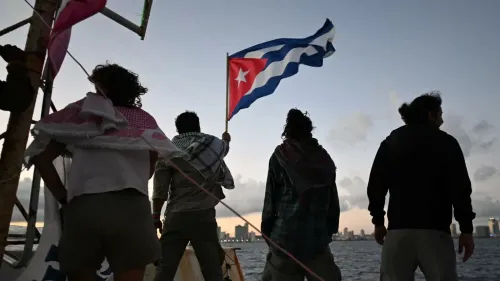 مجموعة من الأشخاص يقفون على ضفاف البحر في هافانا، يحمل أحدهم العلم الكوبي، مع خلفية المدينة في الغسق، تعبيرًا عن التضامن.