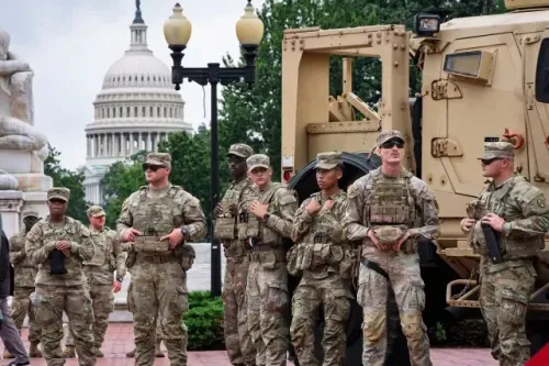 جنود من الحرس الوطني يقفون في وضعية استعداد بالقرب من مبنى الكابيتول الأمريكي، مع وجود مركبة عسكرية خلفهم، في سياق احتجاجات ضد نشر القوات.