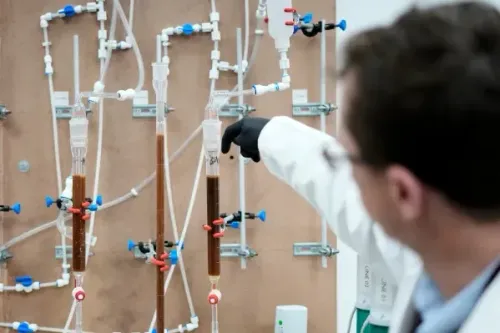 تظهر الصورة باحثًا يرتدي معطفًا مختبريًا، يشير إلى أنابيب زجاجية تحتوي على سوائل ملونة، في مختبر يركز على استخراج العناصر الأرضية النادرة.