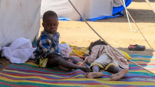 طفل يجلس على سجادة ملونة بجانب طفل آخر مستلقي على الأرض، في سياق أزمة إنسانية في الفاشر بالسودان.