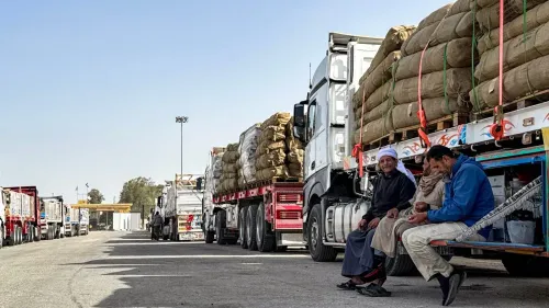 شاحنات محملة بالمساعدات الإنسانية متوقفة عند معبر رفح، مع وجود رجال يجلسون بالقرب منها، استعدادًا لإدخال المساعدات إلى غزة.