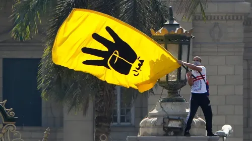 رجل يحمل علم جماعة الإخوان المسلمين الأصفر في ميدان عام، مع خلفية لمبانٍ تاريخية وأشجار، يعبر عن الاحتجاج على التصنيفات الأمريكية.