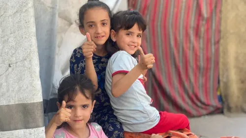 ثلاث فتيات صغيرات يجلسن معًا في مكان مفتوح، يبتسمن ويرفعن أصابعهن بإشارة الإعجاب، في أجواء عيد الفطر بغزة.