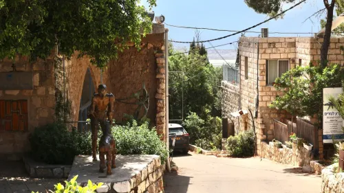 تمثال برونزي لرجل مع كلبه، يقع في قرية عين حوض الحجرية، محاط بمنازل فلسطينية قديمة وأشجار، مع إطلالة على البحر الأبيض المتوسط.