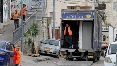 عمال يرتدون سترات برتقالية ينقلون صناديق من شاحنة في حي سلوان، وسط توتر بعد طرد عائلات فلسطينية من منازلها.