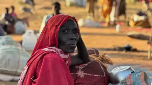 امرأة ترتدي غطاء رأس أحمر، تجلس بين أمتعة في مخيم للنازحين، تعكس معاناتهم في ظل الصراع في السودان.