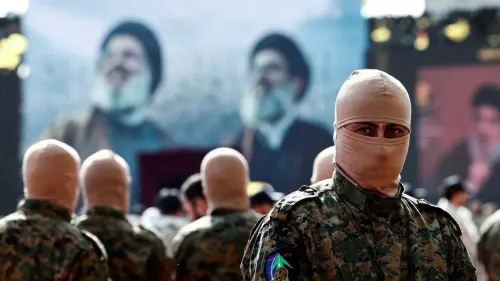 مقاتل من حزب الله يرتدي غطاء رأس كامل، يقف في المقدمة بينما يتواجد آخرون في الخلفية، مع صور لقادة الحزب على الشاشة.