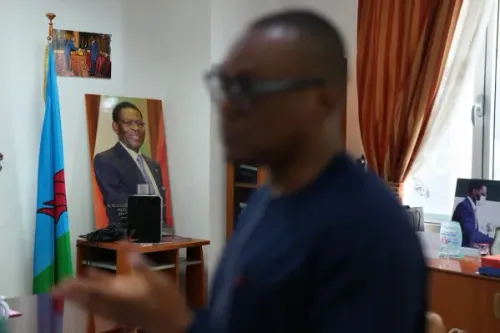 صورة لمكتب في فندق Bamy، يظهر فيها علم غينيا الاستوائية وصورة للرئيس Teodoro Obiang، مع شخص يتحدث في الخلفية.