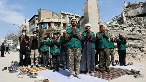 مجموعة من الرجال يرتدون سترات خضراء يؤدون الصلاة في موقع دمار، مع مبانٍ مهدمة في الخلفية، مما يعكس الأوضاع الأمنية المتوترة في إيران.