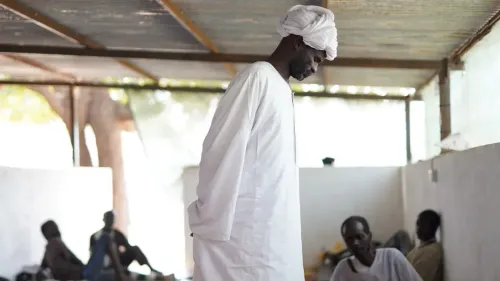 رجل يرتدي ثوبًا أبيض يقف في مكان شبه مظلم، بينما يجلس آخرون في الخلف، ما يعكس الوضع الإنساني الصعب في الفاشر بعد سقوط المدينة.