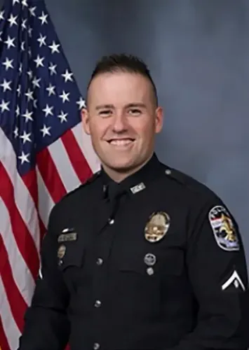 صورة لضابط شرطة مبتسم يرتدي زيًا رسميًا، يقف أمام علم الولايات المتحدة، في سياق قضايا العدالة المتعلقة بمقتل بريونا تايلور.