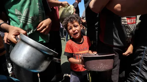 طفل فلسطيني يبكي وهو يحمل وعاءً فارغًا، محاطًا بأشخاص آخرين، مما يعكس معاناة المدنيين في غزة في ظل الظروف الصعبة.