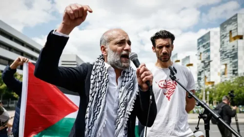 رامي شعث يتحدث في مظاهرة لدعم فلسطين، مرتديًا كوفية فلسطينية، مع وجود علم فلسطيني خلفه، في أجواء احتجاجية.