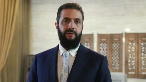 رجل يرتدي بدلة رسمية يتحدث في مؤتمر صحفي، مع خلفية من الزخارف التقليدية، يعكس أهمية الصفقات الاقتصادية في سوريا.