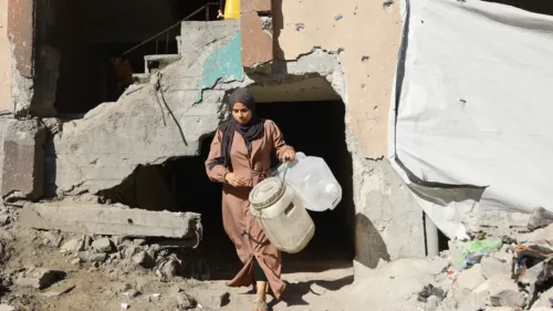 امرأة ترتدي عباءة وتحمل جالونين من الماء، تخرج من مبنى مدمر في غزة، تعكس آثار النزاع المستمر والوضع الإنساني الصعب.