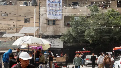 شارع مزدحم في غزة، مع وجود لافتات انتخابية معلقة على المباني، وأشخاص يتجولون تحت مظلة، مما يعكس النشاط الاجتماعي والسياسي في المنطقة.