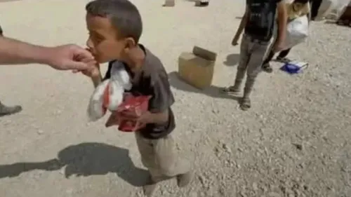 طفل فلسطيني صغير يستلم الطعام من شخص بالغ في موقع توزيع مساعدات، مع أطفال آخرين في الخلفية. تعكس الصورة ظروف الإغاثة الإنسانية في غزة.
